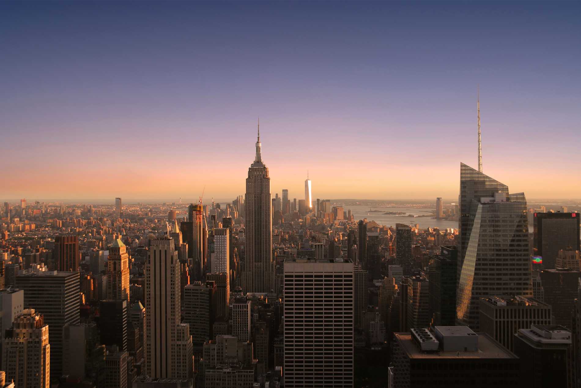 The sun setting over the New York skyline with the Empire State Building in the foreground and One World Trade Center gleaming in the background.