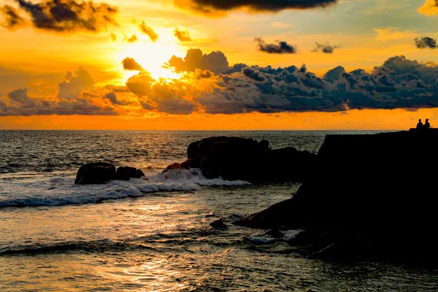 Two people sit on the silhouette of Galle Fort watching the bright orange sun set over the sea.