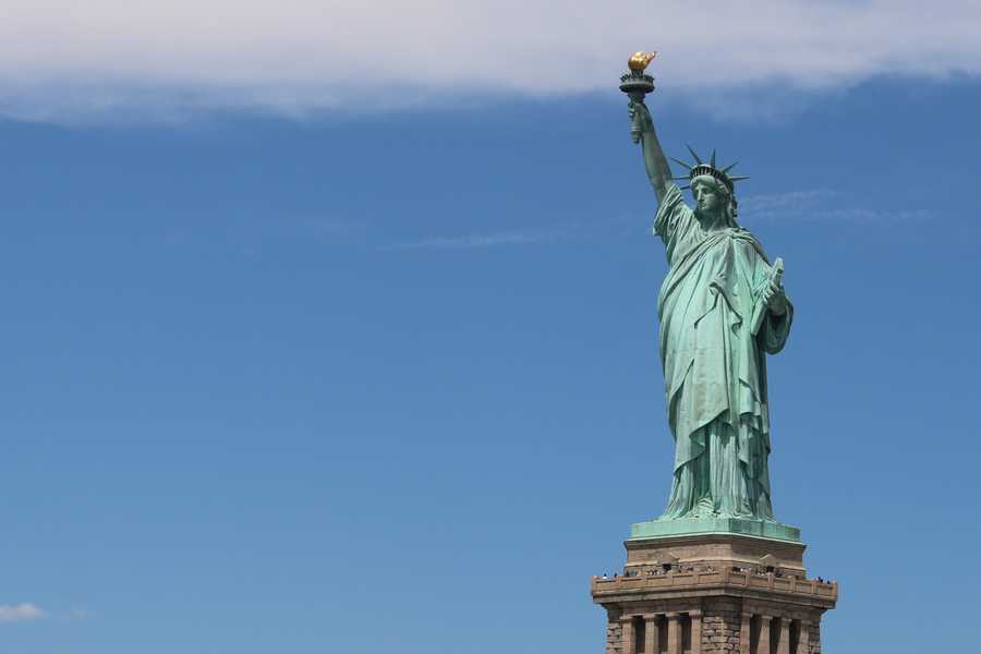 A green Statue of Liberty in front of a bright blue sky holding her torch aloft.